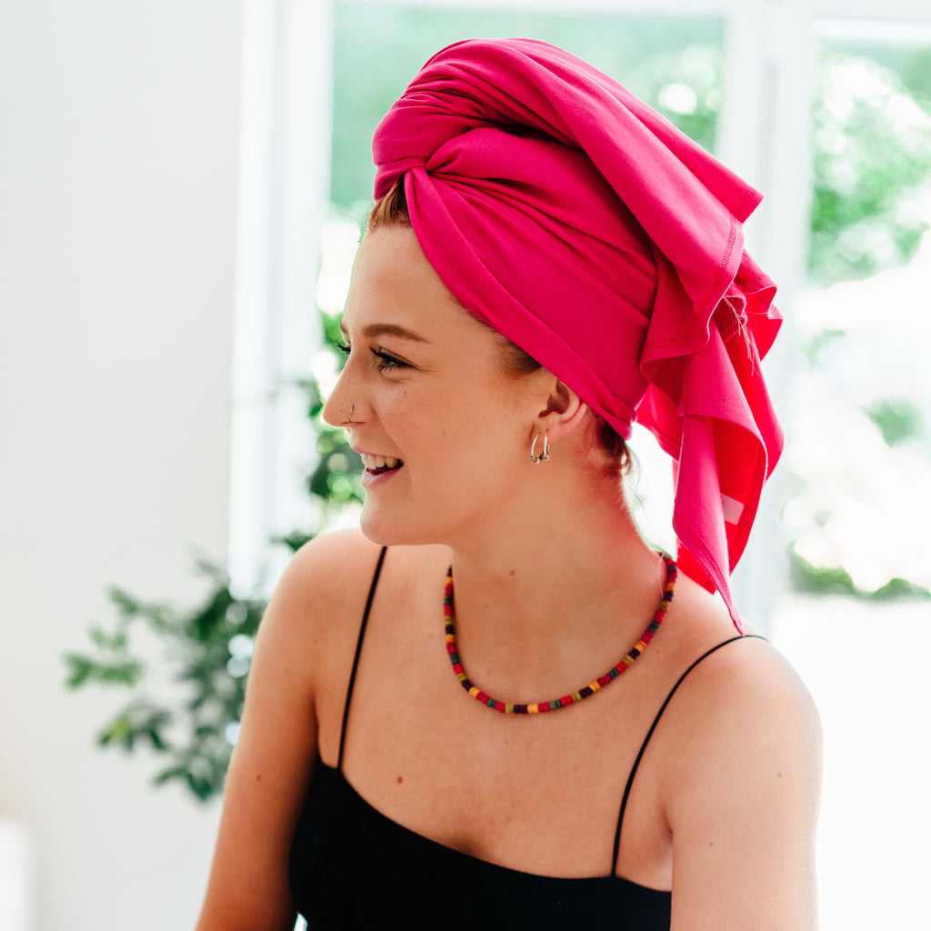 A smiling woman wearing a vibrant pink headwrap and a colorful necklace.