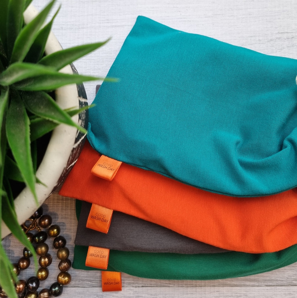 Folded orange and teal Good Wash Day reusable face wipes - pack of 7 + bag - small made of organic cotton with visible labels, beside a plant and a string of beads on a wooden surface.