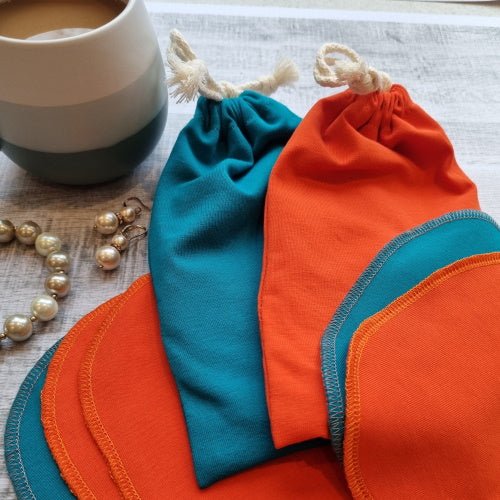 Two Good Wash Day reusable face wipes, one blue and one orange, on a table beside a string of pearls and a cup.
