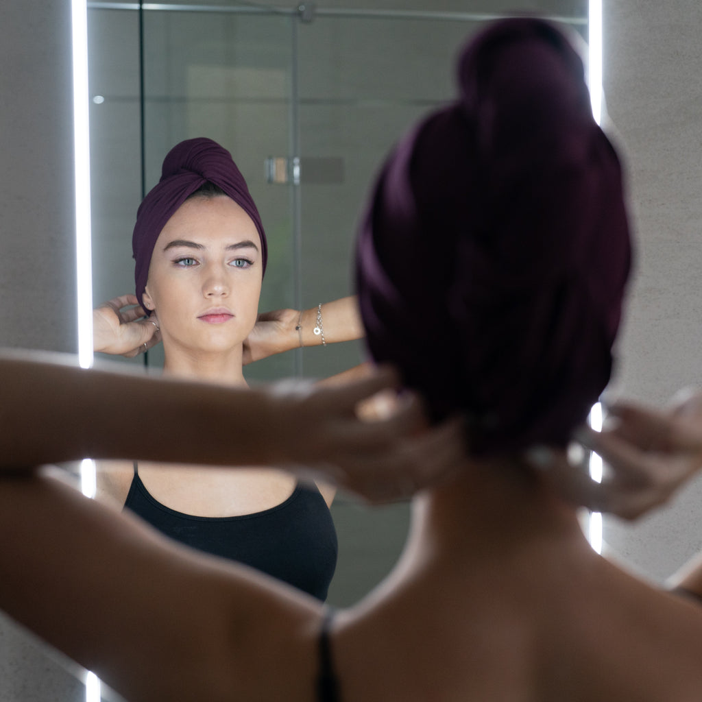 A person, wearing a black top, adjusts The Seconds head wrap made from organic jersey cotton by Good Wash Day while looking at themselves in a well-lit bathroom mirror.