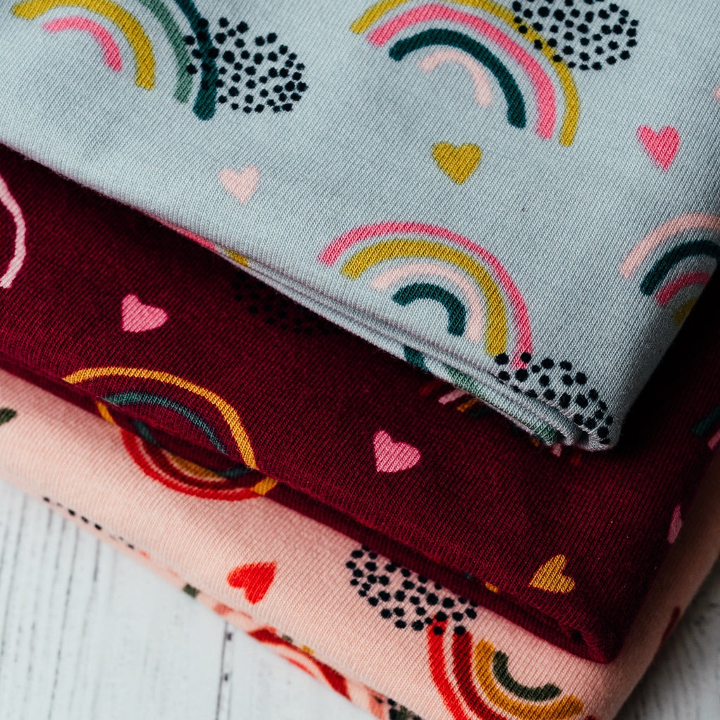 Close-up of three folded pieces of The Rainbows - PRE-ORDER by Good Wash Day stacked on top of each other. Each handmade in the UK, they feature a colorful rainbow and heart pattern on light blue, maroon, and light pink backgrounds. Suitable for sensitive skin, these fabrics add charm to any project.