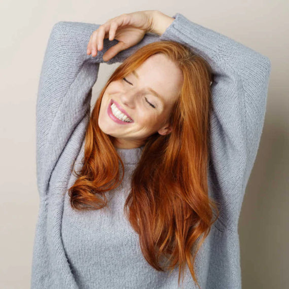 Woman in gray sweater smiling with eyes closed and stretching her arms upwards.