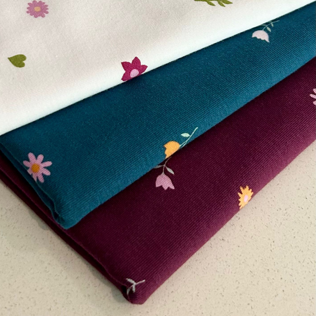 Three stacked fabric pieces: top with small multicolored flowers on organic jersey cotton, middle teal, and bottom burgundy with sparse floral pattern of The Ditsy Prints by Good Wash Day.