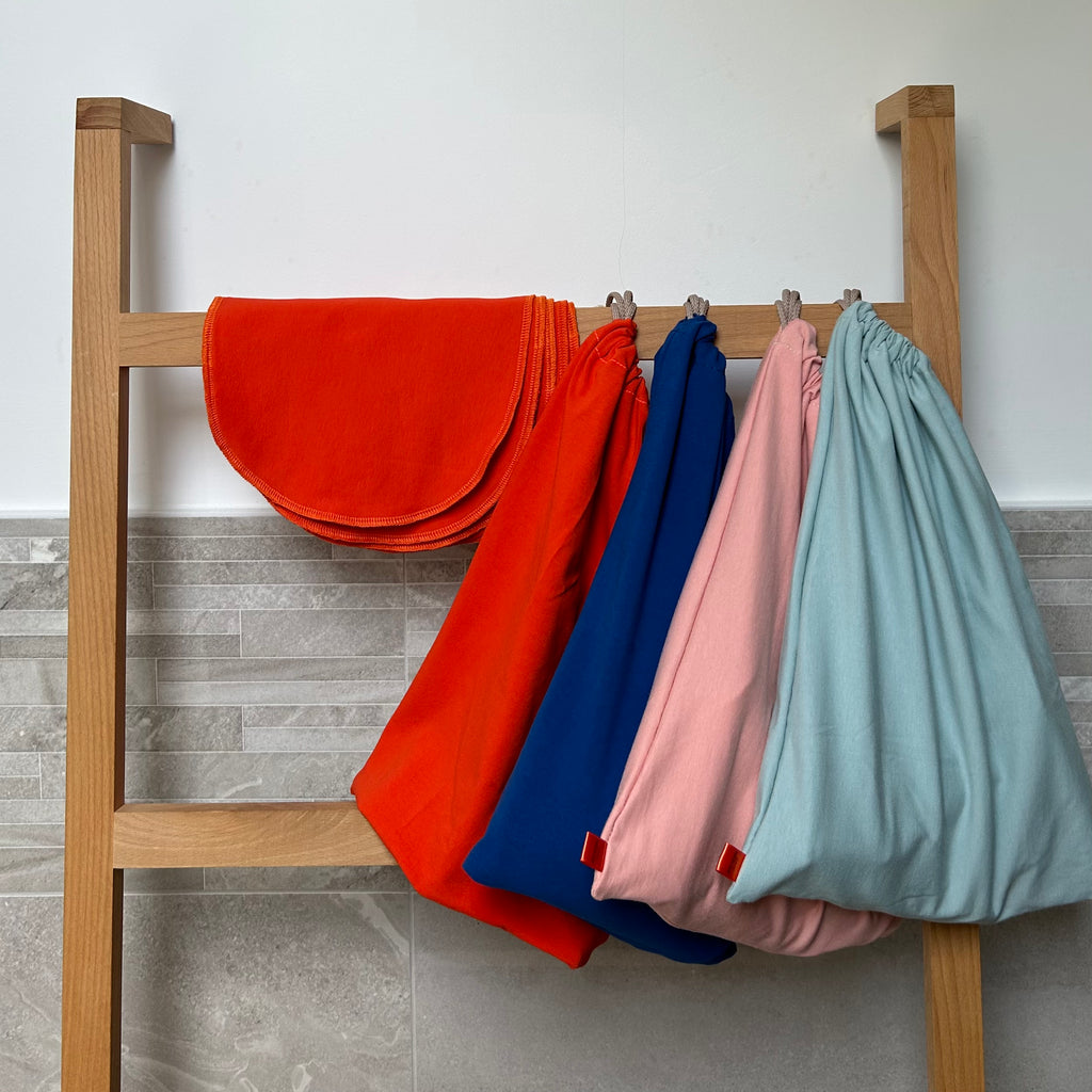 Four colorful Good Wash Day organic cotton reusable face cloths hanging on a wooden rack against a tiled wall, perfect for sensitive skin.