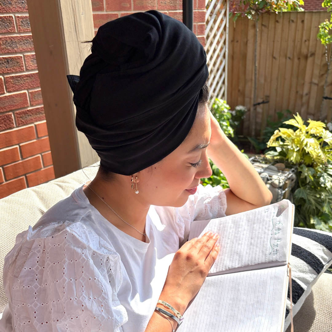 Woman sitting in the garden reading wearing a black organic jersey cotton hair drying towel wrap – Good Wash Day